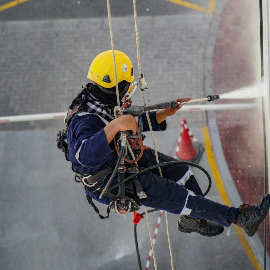 A worker in safety gear high up cleaning a building with a pressure washer in Dubai.